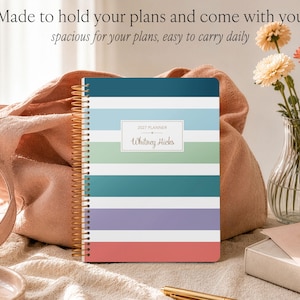 Puede incluir: Una agenda 2027 con encuadernaci&oacute;n en espiral dorada que presenta rayas horizontales en azul oscuro, azul claro, verde, blanco, verde azulado, morado y coral. La portada muestra el texto 2027 PLANNER y el nombre Whitney Hicks en un rect&aacute;ngulo blanco.
