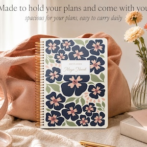 May include: A spiral-bound 2025 planner with a white cover featuring a dark blue and light pink floral pattern and green leaves. The center label reads 2025 PLANNER and Maya Novak. The text above the planner reads Made to hold your plans and come with you, spacious for your plans, easy to carry daily.