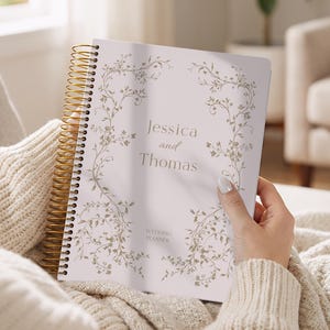 May include: A wedding planner with a gold spiral binding is held open, displaying the names "Jessica and Thomas" in elegant script. The cover features a delicate floral design in a soft, muted color palette.