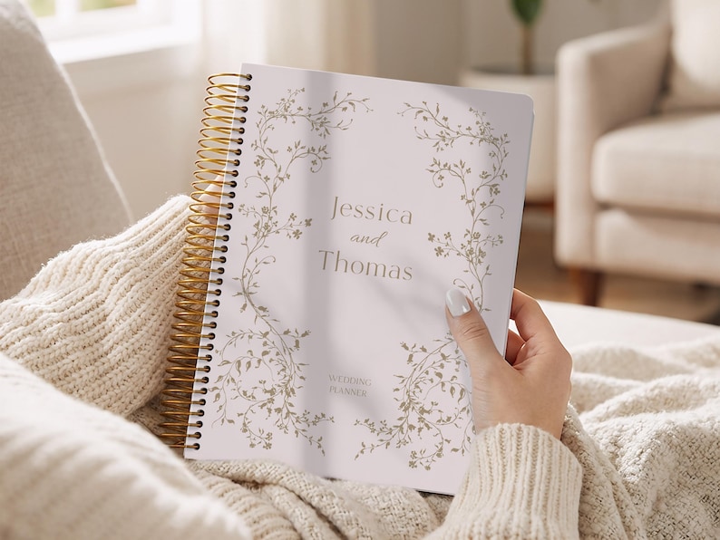May include: A wedding planner with a gold spiral binding is held open, displaying the names "Jessica and Thomas" in elegant script. The cover features a delicate floral design in a soft, muted color palette.