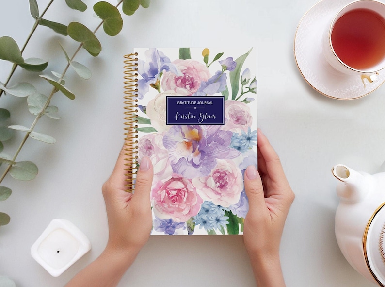 May include: A white spiral bound gratitude journal with a watercolour floral design featuring pink, purple, and blue flowers. The journal cover has the text "Gratitude Journal" and "Karsten Glover" printed in a navy blue rectangle.