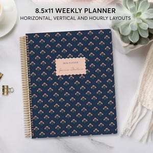 May include: A navy blue 8.5x11 weekly planner with a gold spiral binding. The cover features a repeating diamond pattern with small pink flowers. A central light pink scalloped label displays the text 2026 PLANNER and Jessica Fountain.