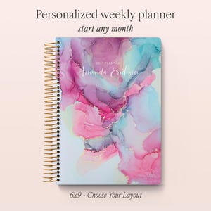 May include: A personalised weekly planner with a gold spiral binding. The cover features an abstract alcohol ink design in shades of pink, purple, and teal with gold accents. The text reads Personalised weekly planner, start any month, 2027 PLANNER, Amanda Eriksen, and 6x9 • Choose Your Layout.