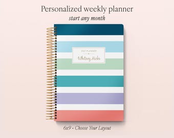 May include: A personalized weekly planner with a gold spiral binding, featuring horizontal stripes in dark blue, light blue, white, green, teal, lavender, and coral. A central white label displays the text 2027 PLANNER and Whitney Hicks.