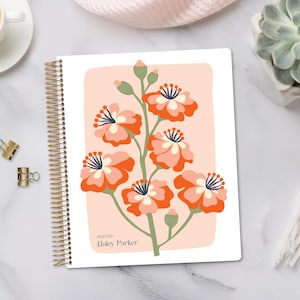 May include: A spiral-bound planner with a floral design. The cover features a branch with orange and white flowers on a peach-colored background. The text "2025-26 Haley Parker" is printed at the bottom. Gold binder clips and a cup of coffee are visible.