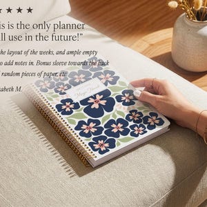 May include: A spiral-bound planner featuring a white cover with a repeating pattern of dark blue flowers with light pink centers and sage green leaves. A central white rectangular label displays the text 2025 PLANNER and Maya Novak.