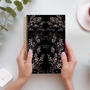 May include: A black gratitude journal with a gold spiral binding. The cover features a floral design with gold and pink accents. The text "Ashley Murray" and "Gratitude Journal" is printed on the cover.