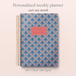 May include: A weekly planner with a blue cover featuring a repeating pattern of small pink flowers and dark blue leaves. The planner has a gold spiral binding and a central pink rectangular label with the text 2027 PLANNER Jessica Fontaine.