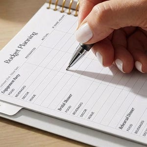 May include: A hand holding a pen writing on a spiral-bound planner. The planner is open to a page titled "Budget Planning" with sections for "Engagement Party," "Bridal Shower," and "Rehearsal Dinner." The page includes columns for "Estimated" and "Actual" costs.