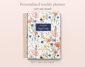 May include: A spiral-bound weekly planner with a white cover featuring a colorful floral pattern of orange, blue, and yellow wildflowers. A dark blue rectangular label in the center displays the text 2027 PLANNER and Kaydee Cretty.