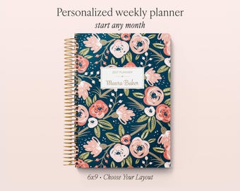 May include: A spiral-bound weekly planner with a dark blue cover featuring a floral pattern of pink and white flowers with green leaves. A central white rectangular label displays the text 2027 PLANNER and Maura Baker. The text Personalized weekly planner start any month and 6x9 Choose Your Layout appear above and below the planner.