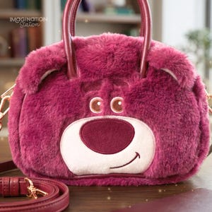 May include: A plush, round, berry-colored handbag with a bear face design. The bag features a white muzzle, a burgundy nose, and the text "CC" above the nose. It has burgundy handles and a detachable shoulder strap. The text "IMAGINATION Station" is visible.
