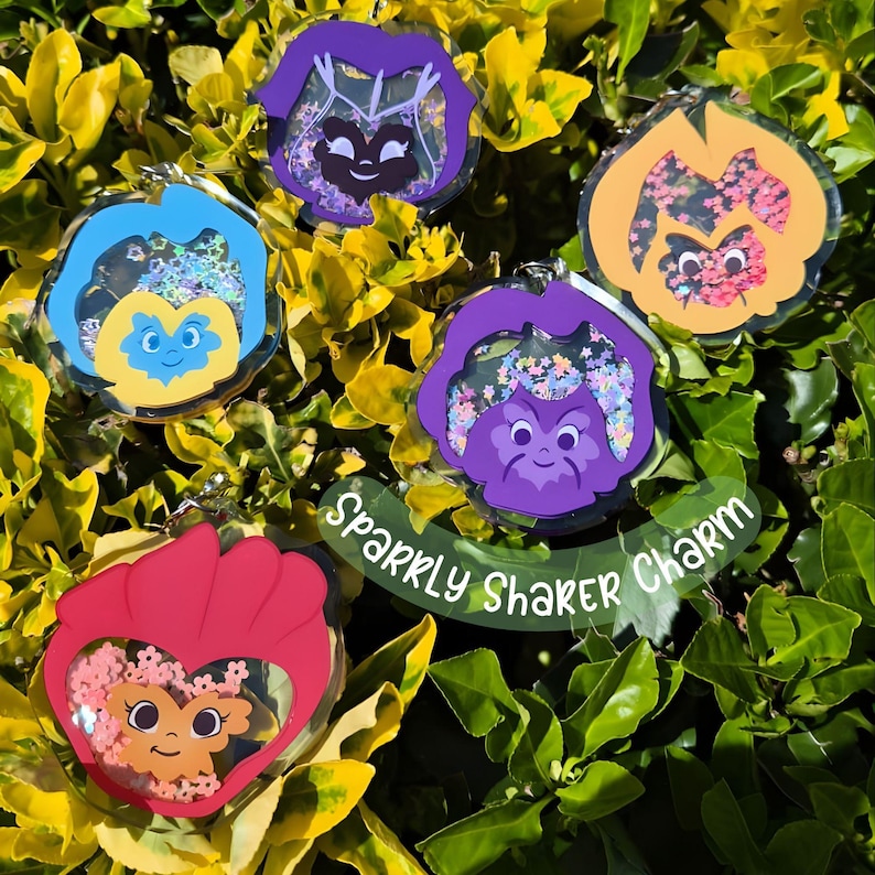 Five colorful acrylic pansy-shaped keychains with smiling faces and glitter, resting on green foliage, with text "Sparkly Shaker Charm."