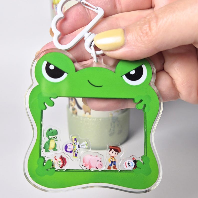 May include: A green frog-shaped keychain with a clear center, featuring cartoon character stickers. The keychain has a white metal clasp. The stickers include a dinosaur, a pig, and a cowboy.