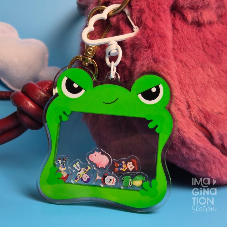 May include: A green frog-shaped keychain with a clear center filled with small character figures. The keychain has a white cloud-shaped clip and a gold-colored clasp. The background is a pink fuzzy texture and a blue surface.