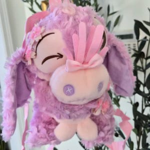 Puede incluir: Un peluche de color lavanda con un dise&ntilde;o caprichoso. Presenta una cara rosa claro con los ojos cerrados, un lazo y flores decorativas. El peluche tiene orejas largas y est&aacute; adornado con cintas, creando una est&eacute;tica suave y juguetona.