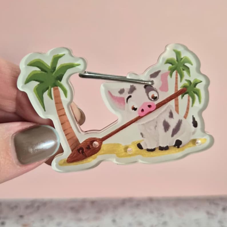 May include: A clear acrylic keychain featuring a cartoon pig character with a brown oar, palm trees, and a sandy beach scene. The pig is white with gray spots and a pink snout. The keychain has a silver metal clasp.