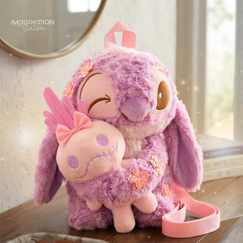 Puede incluir: Una mochila de felpa lavanda con forma de personaje de dibujos animados, gui&ntilde;ando un ojo y abrazando un peluche rosa m&aacute;s peque&ntilde;o. La mochila tiene una correa rosa y est&aacute; decorada con aplicaciones de flores. El texto "IMAGINATION Station" es visible en el fondo.