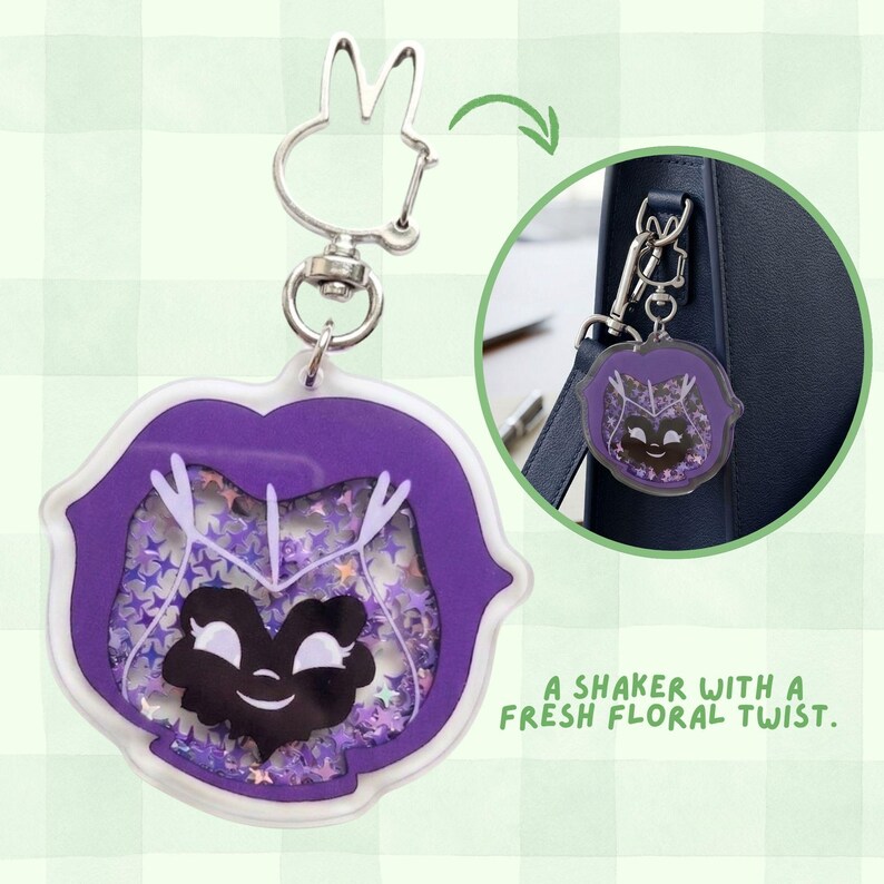A purple acrylic pansy-shaped keychain with a black smiling face and purple star glitter, attached to a silver bunny-ear clip, with an inset showing it clipped to a dark blue bag.