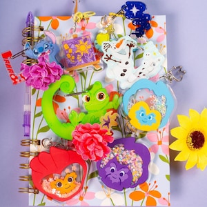 A collection of various cartoon-themed keychains, including several pansy-shaped ones, arranged on an open spiral-bound notebook with a floral pattern, next to a yellow sunflower.