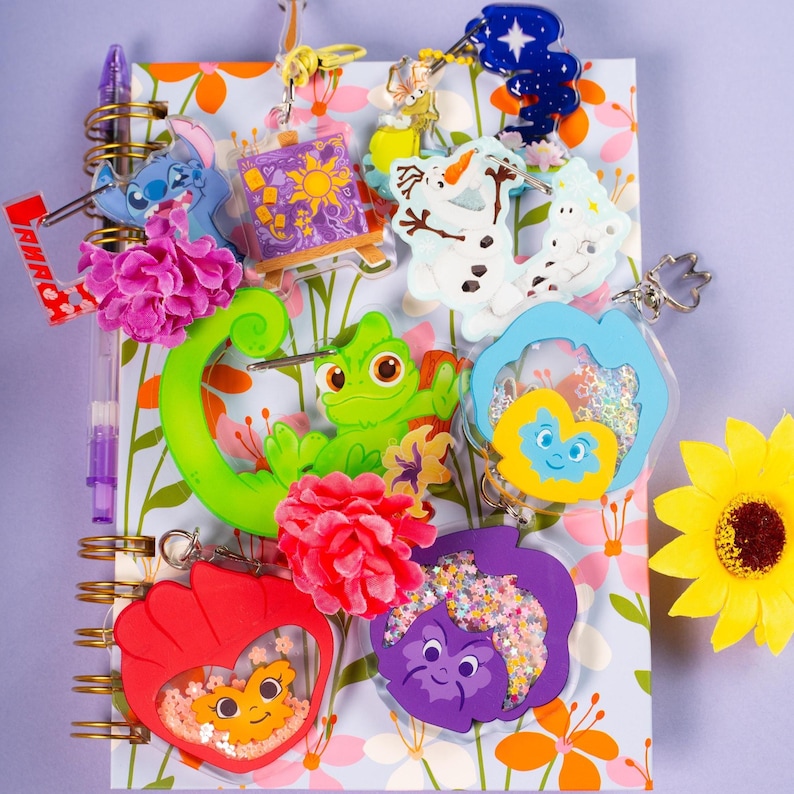 A collection of various cartoon-themed keychains, including several pansy-shaped ones, arranged on an open spiral-bound notebook with a floral pattern, next to a yellow sunflower.