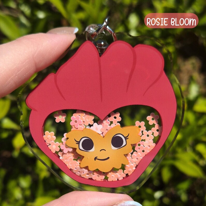 A red acrylic keychain shaped like a pansy with a smiling face, filled with pink flower-shaped glitter, labeled "Rosie Bloom."