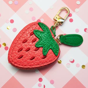 May include: A red strawberry-shaped coin purse with a green leaf design and gold-colored hardware. The purse has a textured surface and a zipper closure. A gold-colored clasp is attached for easy carrying.