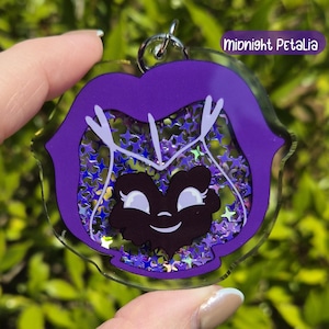 A dark purple acrylic keychain shaped like a pansy with a black smiling face, filled with silver star-shaped glitter, labeled "Midnight Petalia."