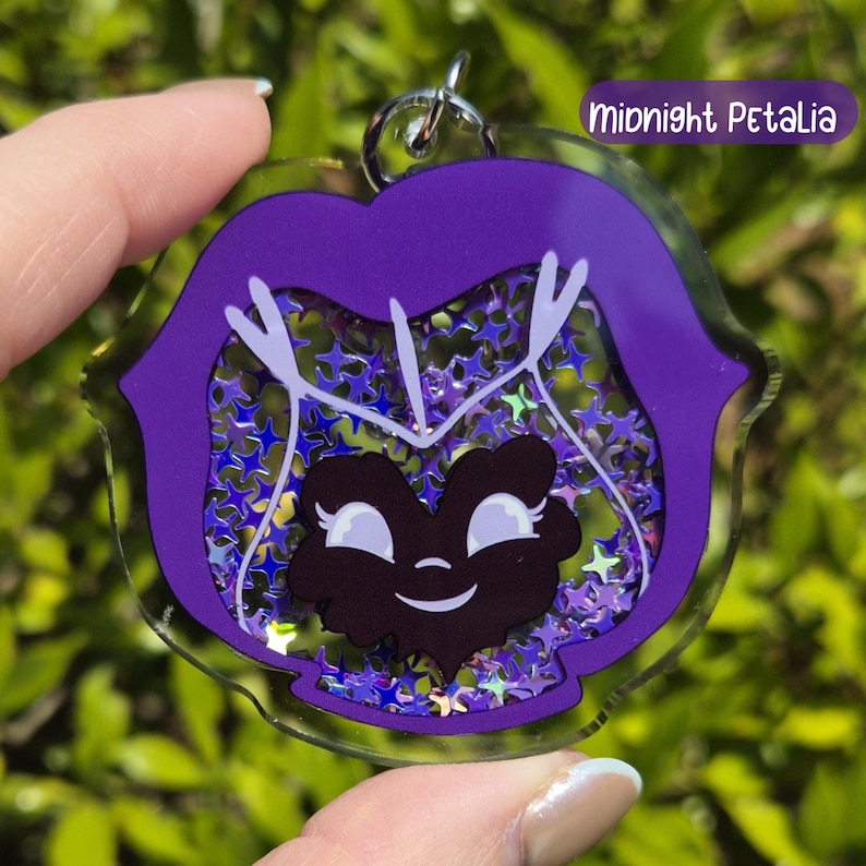 A dark purple acrylic keychain shaped like a pansy with a black smiling face, filled with silver star-shaped glitter, labeled "Midnight Petalia."