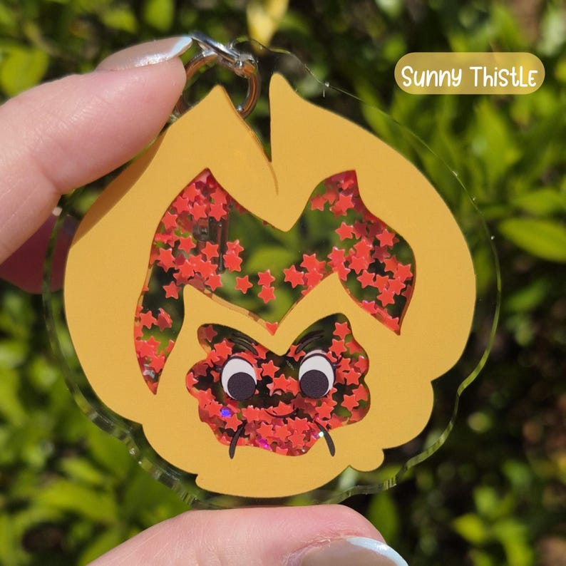 A yellow acrylic keychain shaped like a pansy with a smiling face, filled with red star-shaped glitter, labeled "Sunny Thistle."