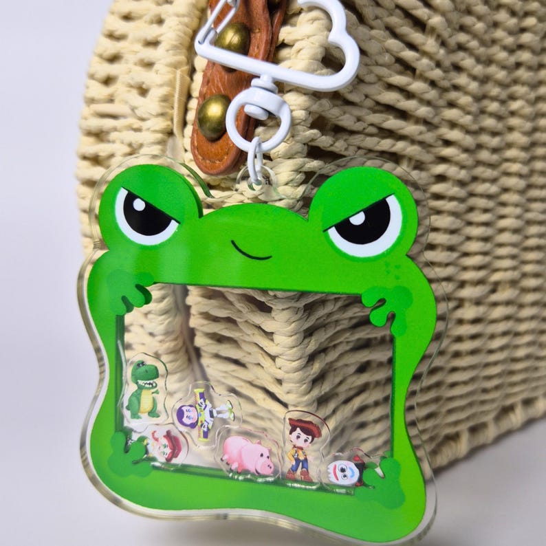 May include: A green frog-shaped keychain with a clear center, featuring small character figures. The keychain has a white metal clasp and is attached to a woven bag. The frog has black eyes and a smiling mouth.