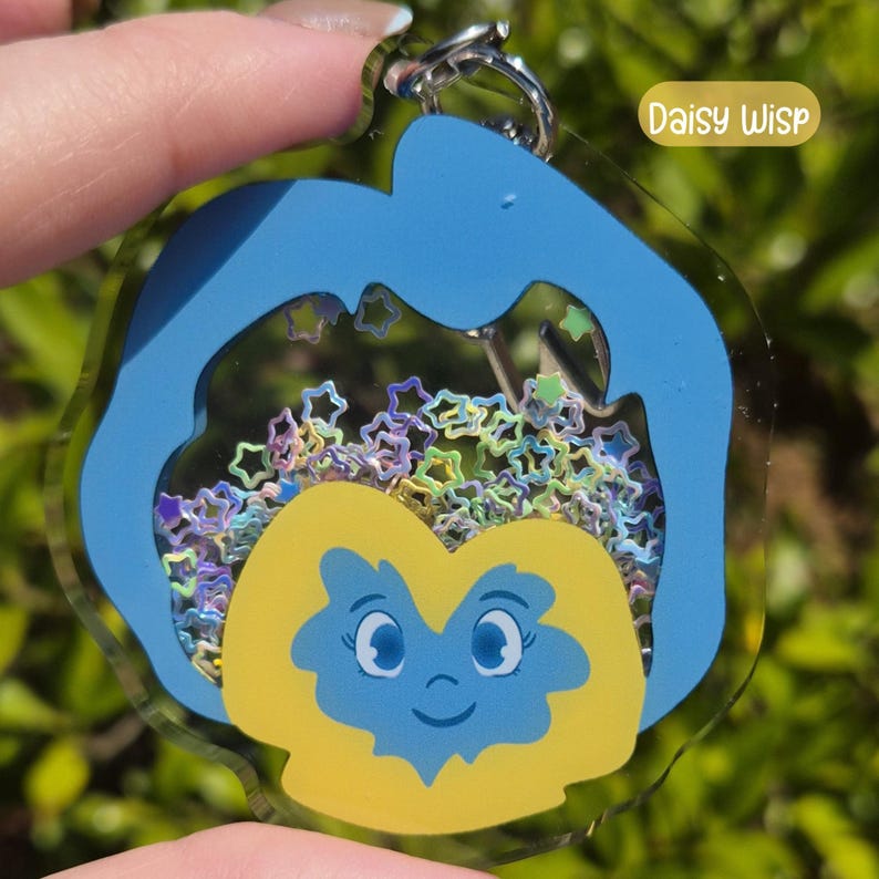 A blue and yellow acrylic keychain shaped like a pansy with a smiling face, filled with colorful star-shaped glitter, labeled "Daisy Wisp."