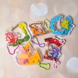 May include: A collection of colorful acrylic keychains featuring cartoon characters. The keychains include a tiger on a magic carpet, a mouse on cheese, a blue fish, a red cat, and a purple insect. Each keychain has a metal ring and a beaded chain.