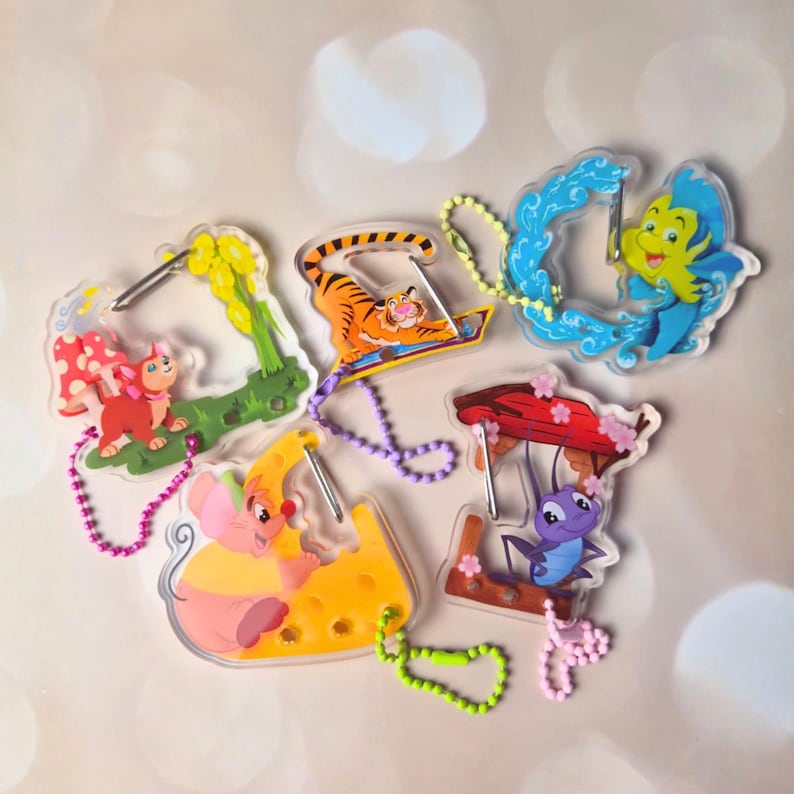 May include: A collection of colorful acrylic keychains featuring cartoon characters. The keychains include a tiger on a magic carpet, a mouse on cheese, a blue fish, a red cat, and a purple insect. Each keychain has a metal ring and a beaded chain.