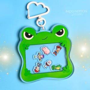 May include: A green frog-shaped keychain with a blue center, featuring cartoon character decals. The keychain has a white cloud-shaped clasp and the text "IMAGINATION Station" on a blue background.