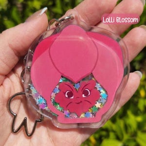 A pink acrylic keychain shaped like a pansy with a smiling face, filled with colorful star-shaped glitter, labeled "Lolli Blossom."