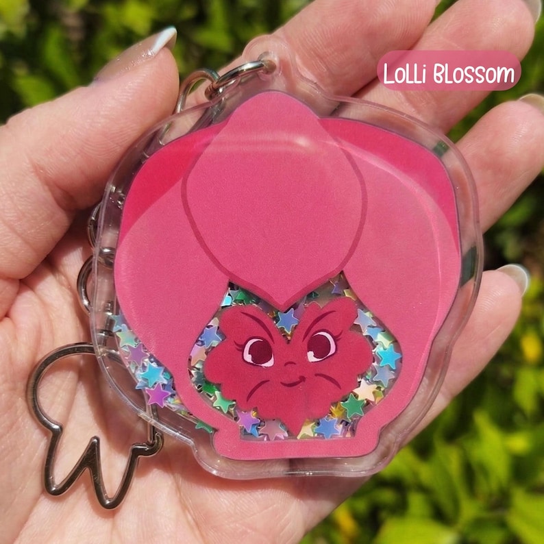A pink acrylic keychain shaped like a pansy with a smiling face, filled with colorful star-shaped glitter, labeled "Lolli Blossom."