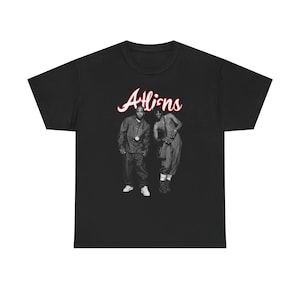 Op de afbeelding: Zwart T-shirt met een zwart-wit afbeelding van twee mensen. Het woord "Aliens" is in een cursief, rood lettertype boven de afbeelding geschreven. Het T-shirt is gemaakt van een zacht, comfortabel materiaal.