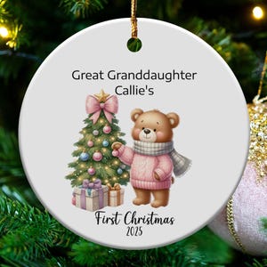 May include: A white, round Christmas ornament with a watercolor illustration of a teddy bear in a pink sweater and scarf, next to a decorated Christmas tree. Text reads "Great Granddaughter Callie's First Christmas 2025".