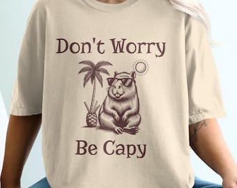May include: Beige t-shirt with the text "Don't Worry Be Capy" and a graphic of a capybara wearing sunglasses, a palm tree, and a sun. The shirt is a relaxed fit.