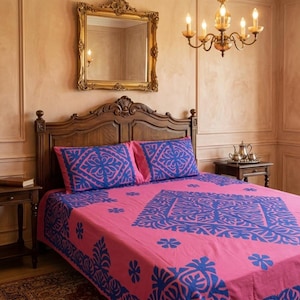 May include: A bedroom scene featuring a bed with a pink and blue patterned bedspread and matching pillowcases. The wooden headboard and side tables complement the warm color palette. A gold-framed mirror and chandelier add elegance.