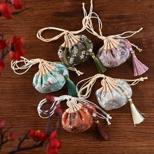May include: Five small, round fabric drawstring pouches in olive green, lavender, teal, silver, and rust orange. Each pouch features a floral pattern, a cream-colored top section, long cream drawstrings, and a matching decorative tassel.