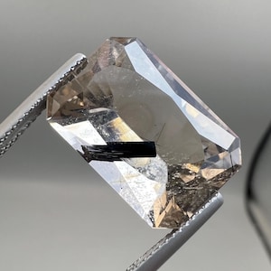 May include: A faceted, rectangular gemstone held by silver tweezers. The gem is a translucent, light brown colour with a dark, black inclusion running through the centre. The gem has many facets that reflect light.