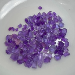May include: A scattering of small, raw amethyst crystals in shades of purple and violet. The crystals are displayed on a white surface, highlighting their natural, irregular shapes and translucent appearance. These gemstones are suitable for jewellery making or collecting.