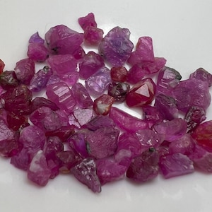 Peut inclure: Un ensemble de pierres précieuses de rubis brutes, non taillées, de différentes nuances de rose et de rouge. Les pierres sont disposées sur une surface blanche, révélant leurs formes naturelles irrégulières et leurs structures cristallines. Les rubis varient en taille et en intensité de couleur.