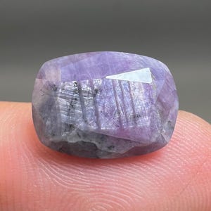May include: A cushion-cut lavender and grey gemstone with a faceted surface. The stone displays natural internal inclusions, visible striations, and a translucent quality with varying shades of purple and dark grey throughout the crystalline structure.