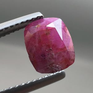 May include: A faceted, reddish-pink gemstone held by a pair of metal tweezers. The gem has a rectangular shape with multiple facets that reflect light. The stone's color varies from deep pink to lighter shades, with some internal inclusions visible.
