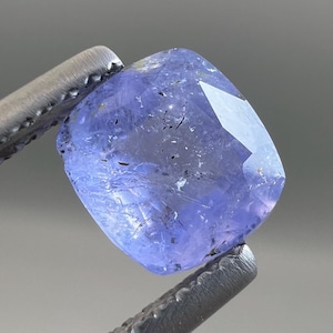 May include: A faceted, square-cut, blue-violet gemstone held by a pair of silver tweezers. The gemstone has a polished, reflective surface with visible internal inclusions. The color is a soft, medium tone, with a slight variation in hue.