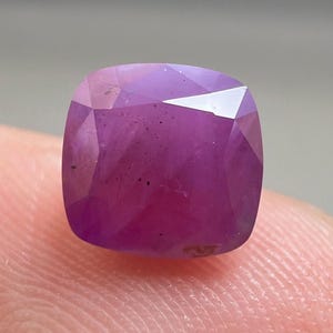 May include: A faceted, cushion-cut gemstone in a deep magenta colour. The gem has a complex internal structure and a polished surface, reflecting light. The stone is held by a finger, showcasing its size and colour.