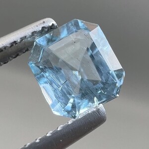 Puede incluir: Una gema azul claro facetada, sujeta con pinzas plateadas. La gema tiene un corte esmeralda con múltiples facetas que reflejan la luz. El color es azul pálido y transparente, con una ligera textura interna. La forma general es rectangular.